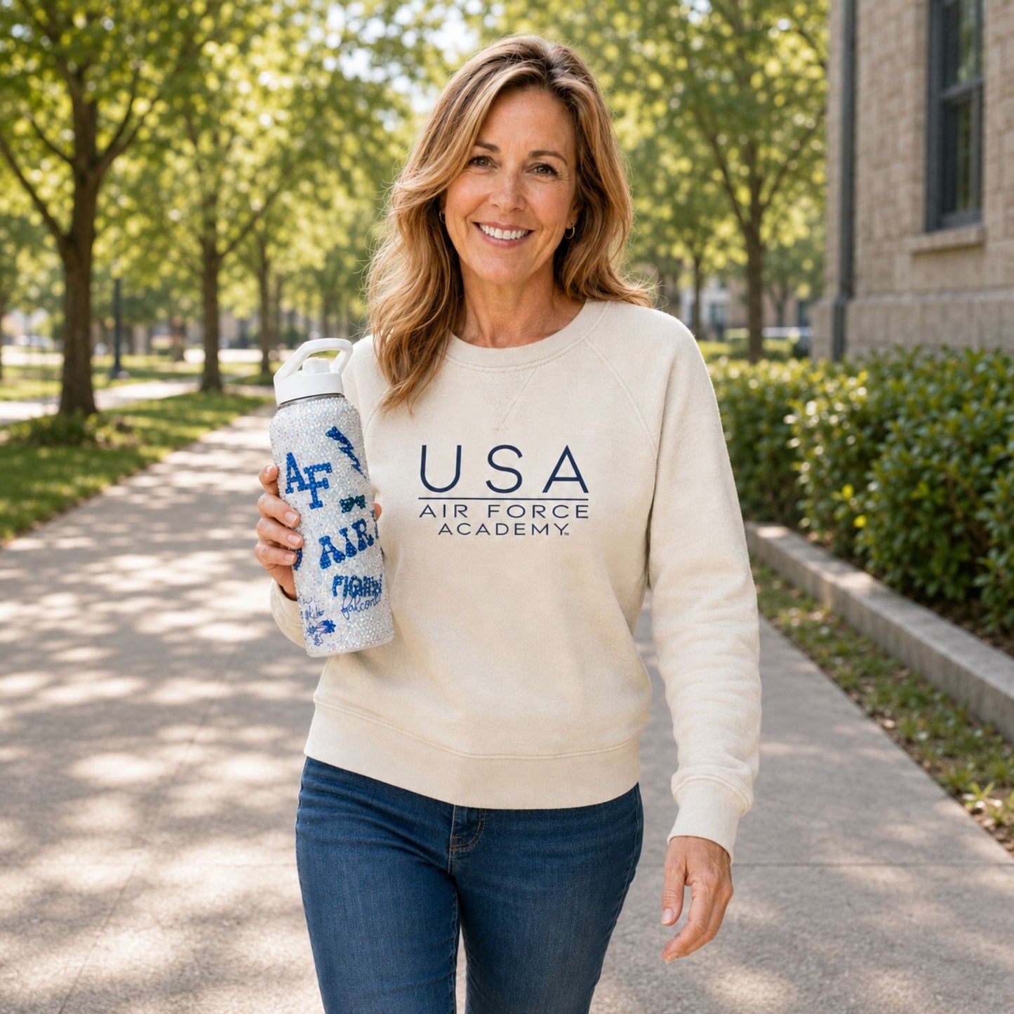 Woman walking outdoors in a cream USA Air Force Academy sweatshirt while holding a rhinestone-covered water bottle.