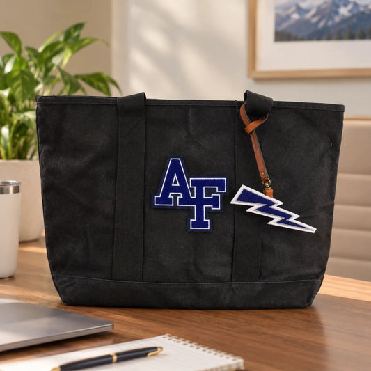 Air Force Lightning Bolt Chenille Bag Charm hanging from a black AF canvas tote on a wooden office desk, featuring a brown leather strap and navy blue lightning bolt design.