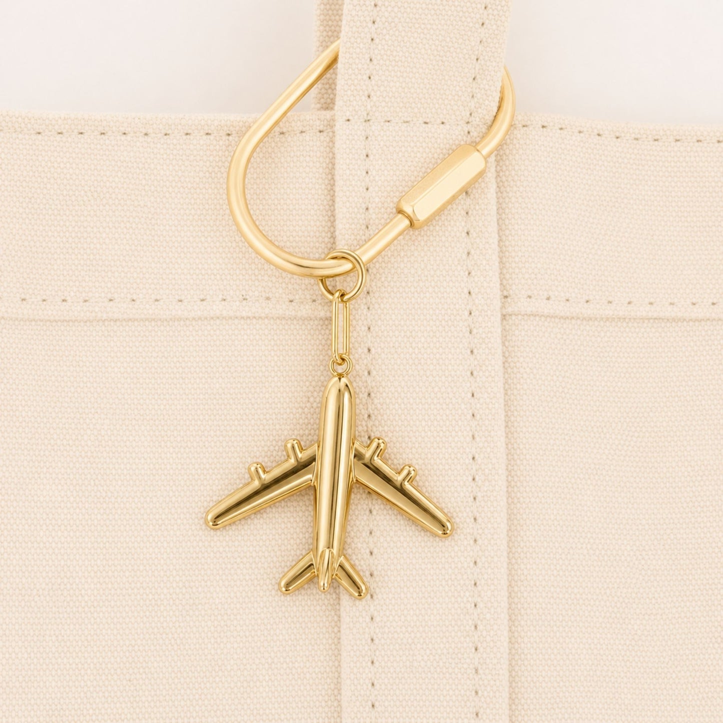 Gold airplane bag charm attached to the handle of a cream canvas tote.