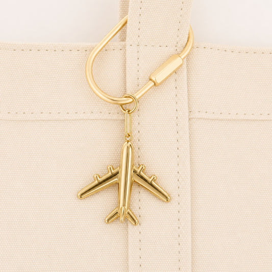 Gold airplane bag charm attached to the handle of a cream canvas tote.