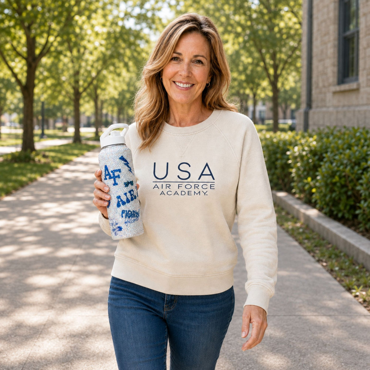 Woman walking outdoors in a cream USA Air Force Academy sweatshirt while holding a rhinestone-covered water bottle.