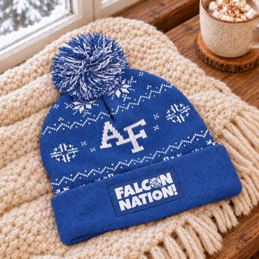 Blue Air Force Falcon Nation beanie resting on a cream knit blanket beside hot cocoa and a window overlooking snowy trees.