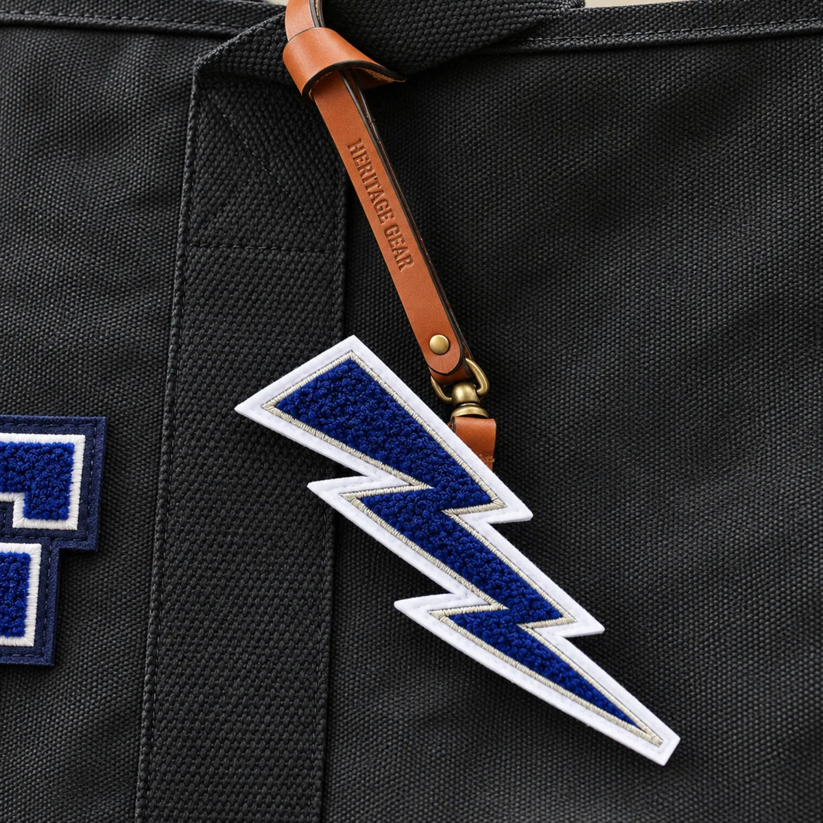 Close-up of the Air Force Lightning Bolt Chenille Bag Charm attached to a black canvas tote, highlighting the textured navy chenille patch, white embroidered border, and embossed Heritage Gear leather strap.