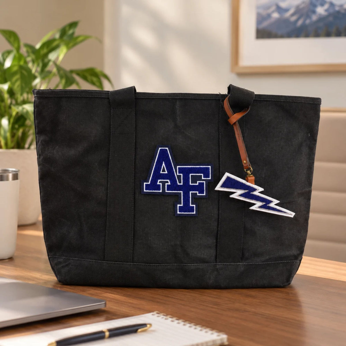 Air Force Lightning Bolt Chenille Bag Charm hanging from a black AF canvas tote on a wooden office desk, featuring a brown leather strap and navy blue lightning bolt design.