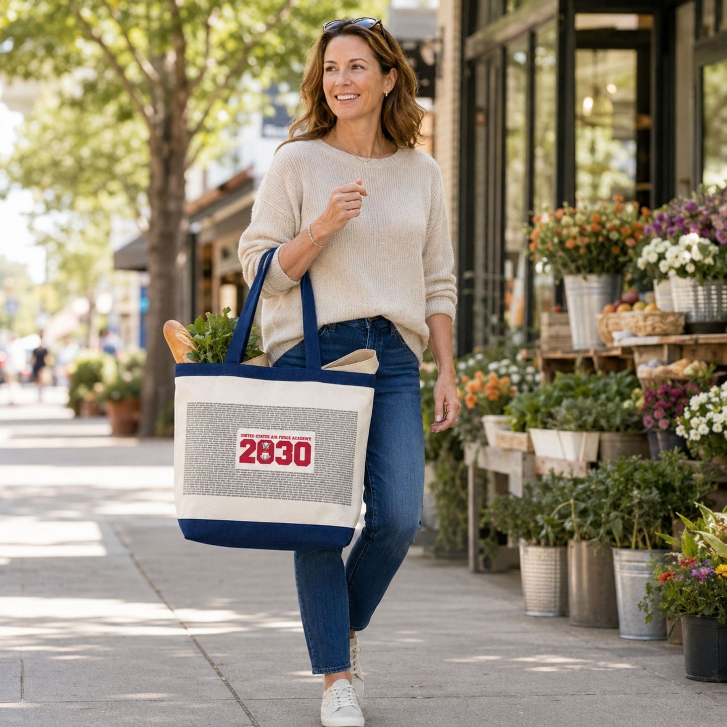 Woman carrying a United States Air Force Academy Class of 2030 tote filled with groceries while walking past a neighborhood flower market.
