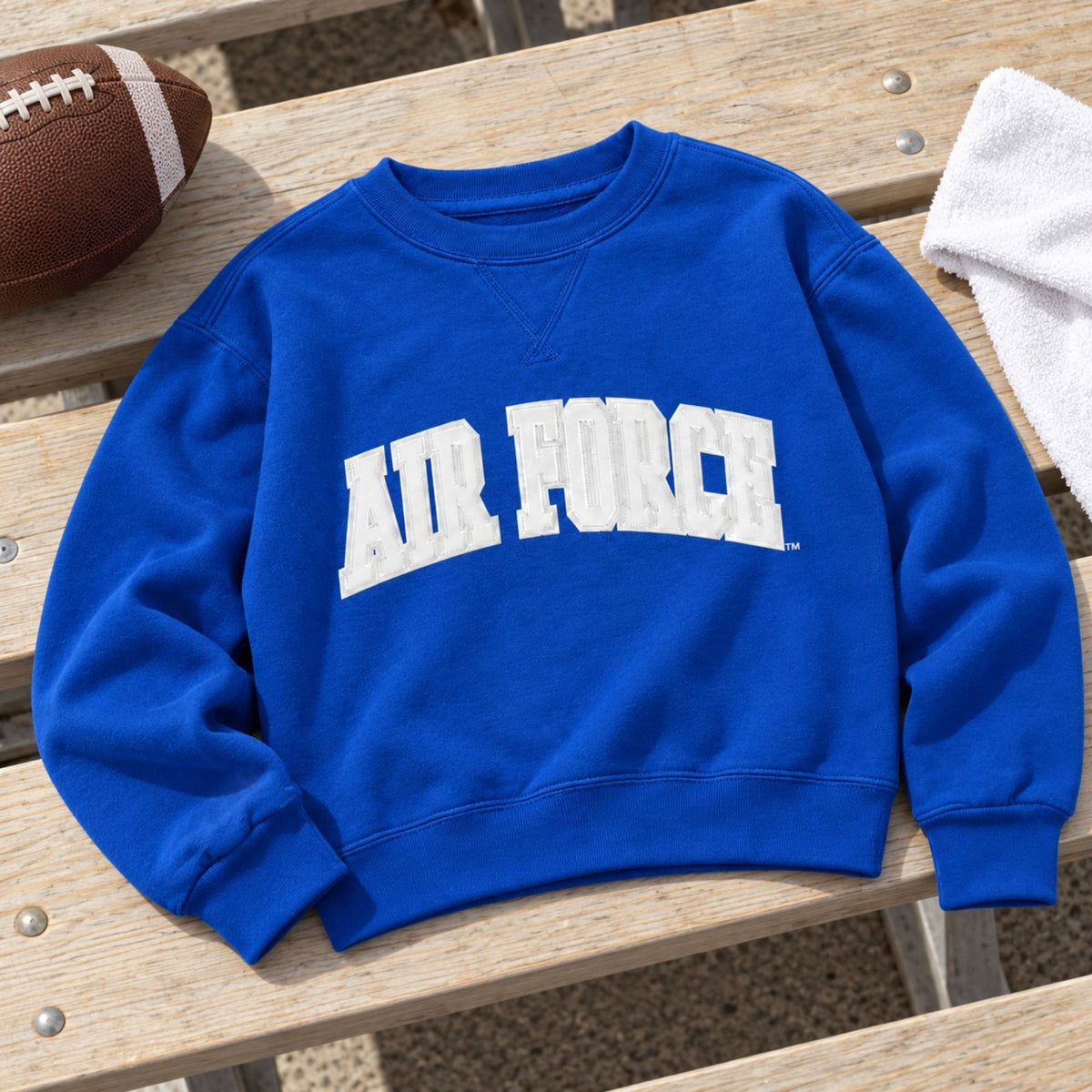 Blue Youth Air Force Courtside Crewneck laid on wooden bleachers beside a football and white rally towel